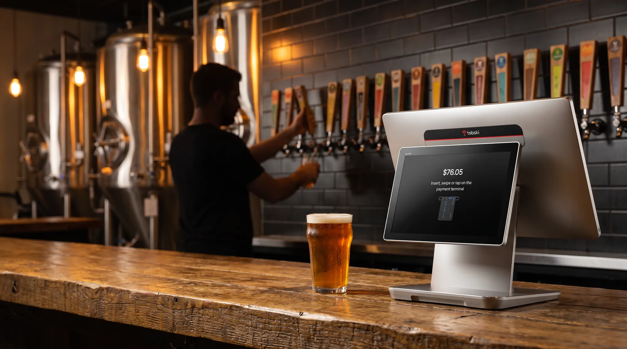 Tabski POS terminal on a brewery bar next to a pint of beer, with taps and fermenters behind