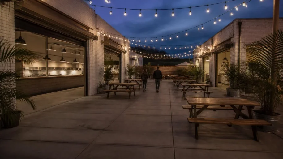 Food hall courtyard at dusk with string lights, open garage doors on brick buildings and wooden picnic tables (illustrative image)