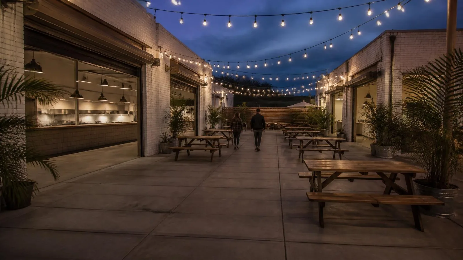 Food hall courtyard at dusk with string lights, open garage doors on brick buildings and wooden picnic tables (illustrative image)