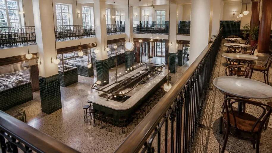Two-level food hall seen from a balcony, with dark green tiled counters, a long central bar, terrazzo floors and globe pendant lights (illustrative image)