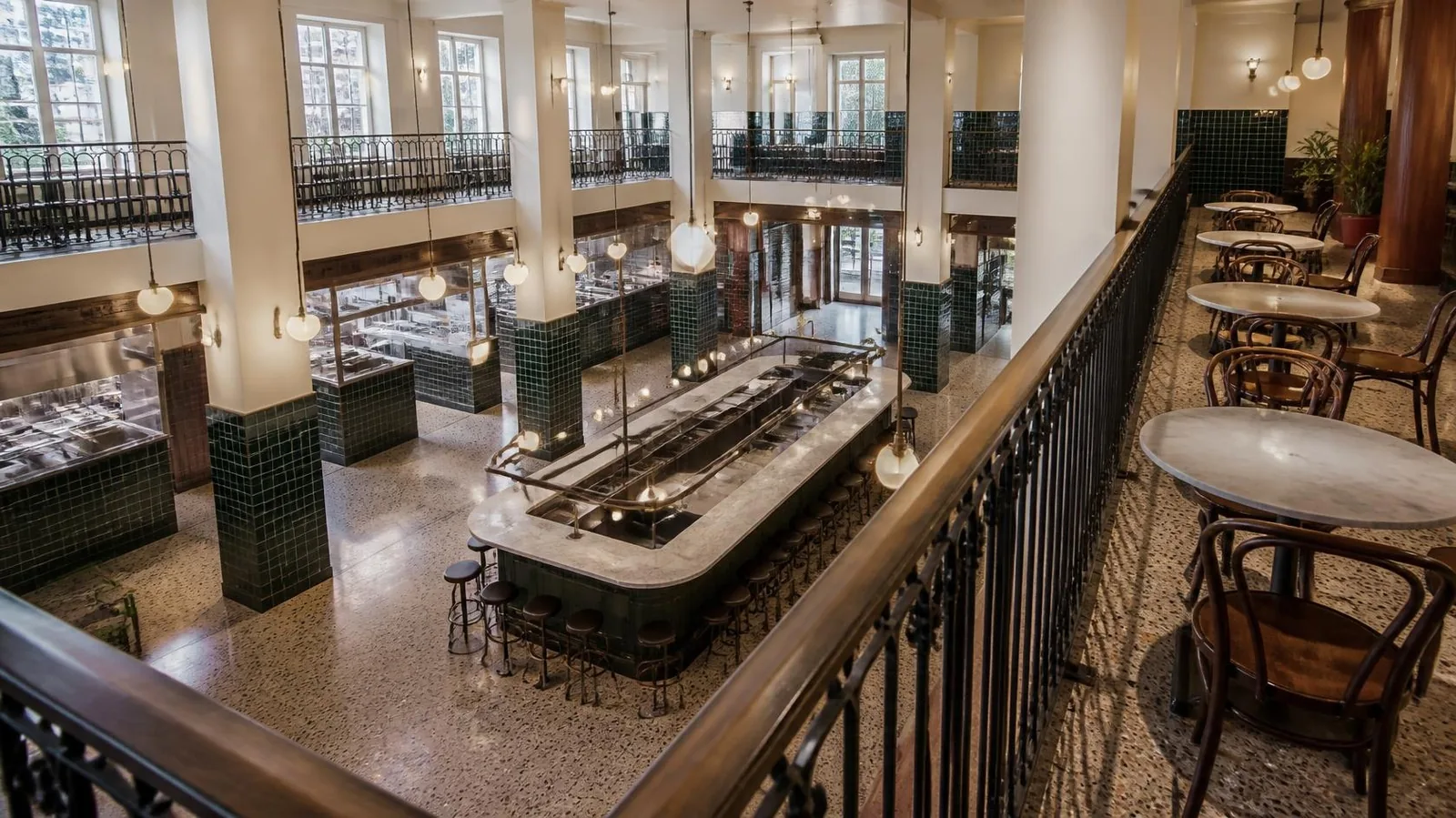 Two-level food hall seen from a balcony, with dark green tiled counters, a long central bar, terrazzo floors and globe pendant lights (illustrative image)